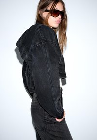 Ženska, oblečena v prekrajšano črno denim jakno in hlače, s sončnimi očali, pozira sstran ob beli ozadju.