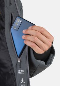 Wasserabweisende Jacke mit einer grauen Außenschicht, die eine Reißverschlusstasche enthält, in der sich ein blauer Smartphone befindet. Auf der Jacke ist die Wasserdichtigkeitsbewertung vermerkt.