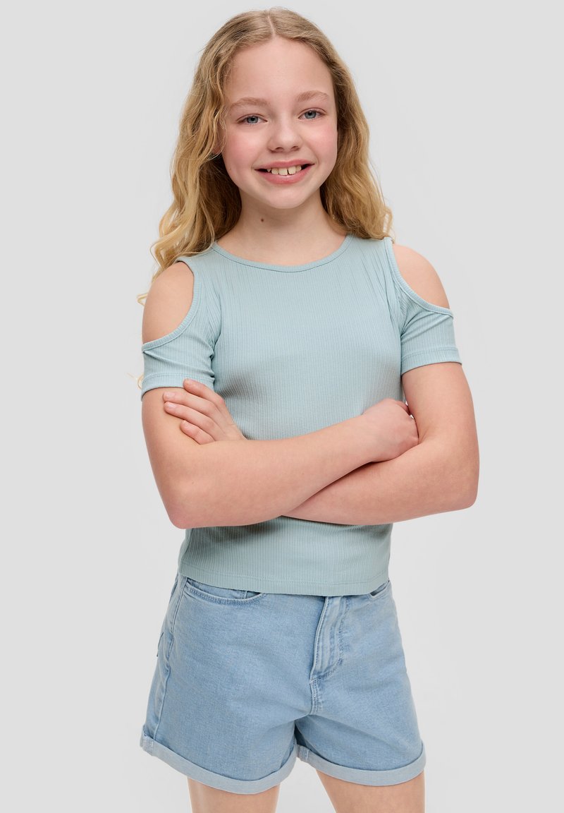 Top court côtelé bleu clair avec découpes épaules nues, associé à un short en denim clair. Le mannequin a des cheveux ondulés et les bras croisés.