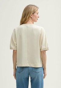 Cremfarbiges, kurzärmliges Oberteil mit lockerer Passform und strukturierten Bündchen. Kombiniert mit hellblauen Jeans, die über Gesäßtaschen und Nähte verfügen.
