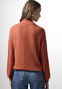 Top a maglia color ruggine con una texture morbida, caratterizzato da un collo alto, maniche lunghe e una vestibilità rilassata, abbinato a jeans in denim blu.