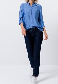 Camisa de botones de color azul claro con un bolsillo en el pecho, combinada con jeans ajustados de mezclilla oscura y zapatillas blancas. Tejido suave con mangas arremangadas.