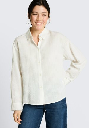 Uśmiechnięta kobieta o ciemnych włosach, ubrana w fakturowaną białą koszulę zapinaną na guziki i niebieskie dżinsy, pozująca z jedną ręką na biodrze.