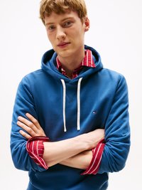 Sudadera azul con cordones blancos, usada sobre una camisa de cuadros roja y blanca. El modelo tiene los brazos cruzados, mostrando las capas de la ropa.