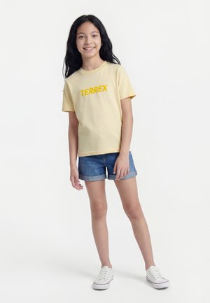 Smilende pige står iført gul TERREX t-shirt, blå denimshorts og hvide sneakers mod en hvid baggrund.