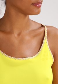 Ärmlös gul topp med rund halsringning som har en flerfärgad mönstrad kant. Slät textur med en lätt lyster.