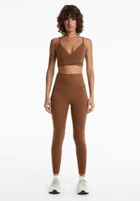 Crop top de sport marron à fines bretelles associé à un legging taille haute assorti. Tissu doux et extensible avec un design sans couture. Baskets blanches.