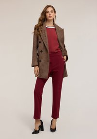 Hnědý houndstooth dvouřadý kabát, vínová hedvábná blůza, vínové kalhoty s bočními knoflíky, černé vysoké podpatky, hladká textura.
