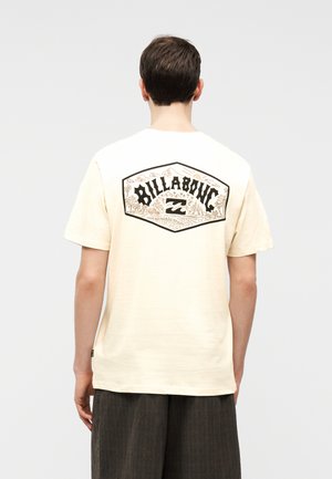 Fiatal férfi krémszínű Billabong pólóban, amelynek hátulján fekete és bézs trópusi logó található, sötét, laza nadrággal párosítva.