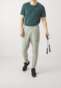 Zielona koszulka z krótkim rękawem, szare spodnie dresowe z elastycznym pasem oraz czarne buty sportowe. Trzyma skakankę. Logo Nike na odzieży.