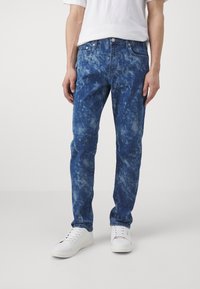 Modré džínové kalhoty s efektem rozstříkaného kyselého praní. Přiléhavý střih, pět kapes, zapínání na kovový knoflík a nošené s bílými teniskami.