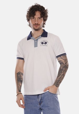 Uomo con capelli ricci, indossa una polo bianca con colletto blu navy e jeans blu, mostra tatuaggi dettagliati su entrambe le braccia, mano in tasca.