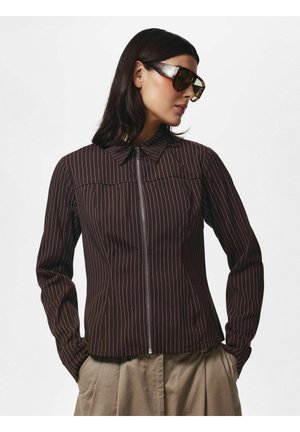 Frau mit schulterlangem dunklem Haar, die große Schildpatt-Sonnenbrille, eine braune gestreifte Zip-up-Jacke und beige plissierte Hosen trägt.