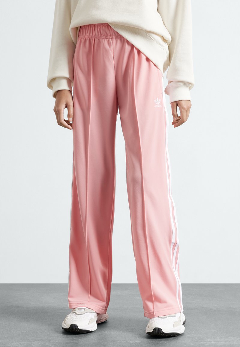 Różowe spodenki sportowe z poliestru z białymi paskami po bokach i elastycznym pasem. Materiał ma gładką fakturę, w połączeniu z białymi trampkami.