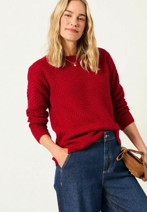 Roter strukturierten Strickpullover mit Rundhalsausschnitt und gerippten Bündchen, kombiniert mit dunklen Jeans. Eine kleine braune Tasche wird von dem Modell gehalten.