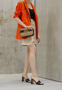 Blazer arancione, top nero, gonna beige e tacchi aperti neri. Una borsa a tracolla con motivo e dettagli dorati è appesa alla spalla.