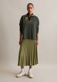 Donna in piedi che indossa una maglietta a maniche lunghe verde scuro, una gonna midi plissettata di colore oliva chiaro, sneaker beige, calzini bianchi e una sciarpa fantasia annodata attorno al collo.