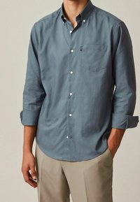 Chemise à boutons pour hommes en coton bleu ardoise. Comprend un col pointu, des manches longues avec poignets roulés et une poche poitrine.