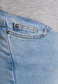 Jeans de ganga azul claro com um design clássico de cinco bolsos. Apresentam costura em cobre e rebites de metal pretos, exibindo um acabamento texturizado.