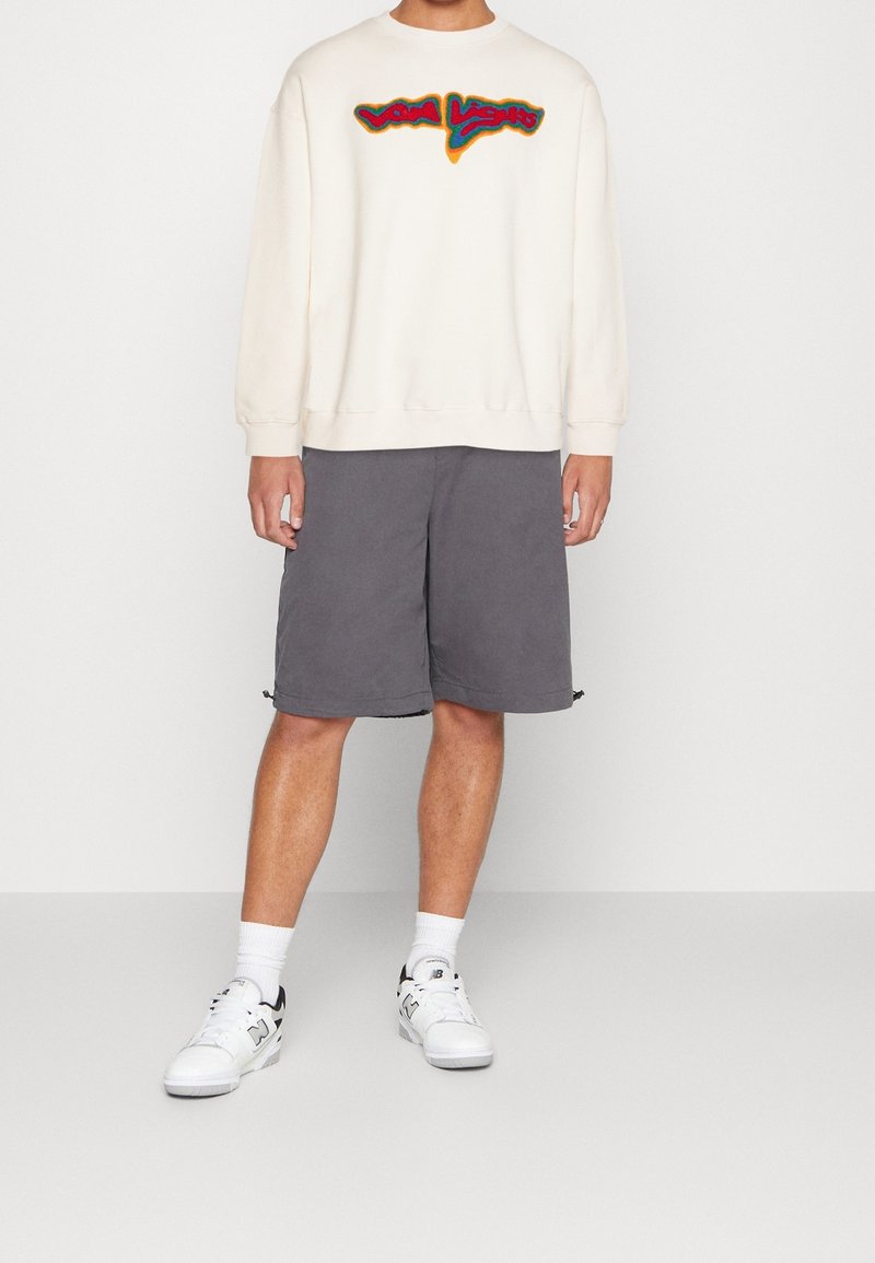 Persona che indossa una felpa bianca con testo colorato, pantaloni corti grigi fino al ginocchio, calze bianche alla caviglia e sneakers bianche con accenti neri.