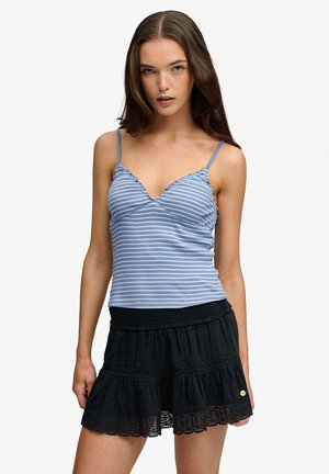 Femme portant un haut sans manches rayé bleu et blanc et une jupe noire à volants avec bordure en dentelle, debout devant un fond uni.