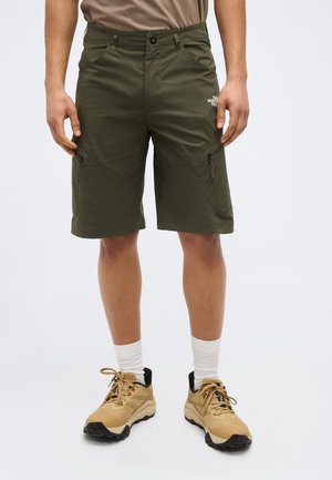 Muž oblečený v olivovo zelených cargo šortkách so zipsovými vreckami, béžových turistických topánkach, bielych ponožkách a hnedom tričku na jednoduchom pozadí.