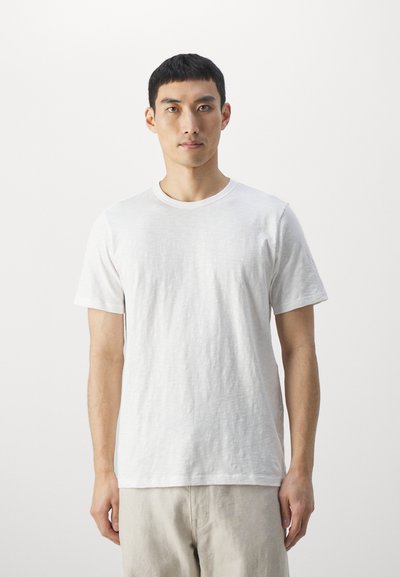 Giovane uomo in piedi di fronte, indossando una semplice maglietta bianca a maniche corte e pantaloni beige chiaro su uno sfondo bianco.