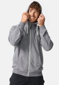 Hanorac gri cu fermoar, buzunar frontal, glugă cu șnur și manșete și tiv striate. Textură moale a țesăturii și detaliu neted al fermoarului.