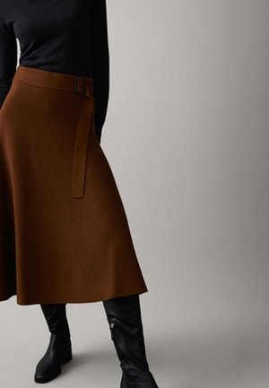 Vrouw draagt een bruine midirok met riem, zwarte top en zwarte kniehoge laarzen, staand tegen een effen lichte achtergrond.