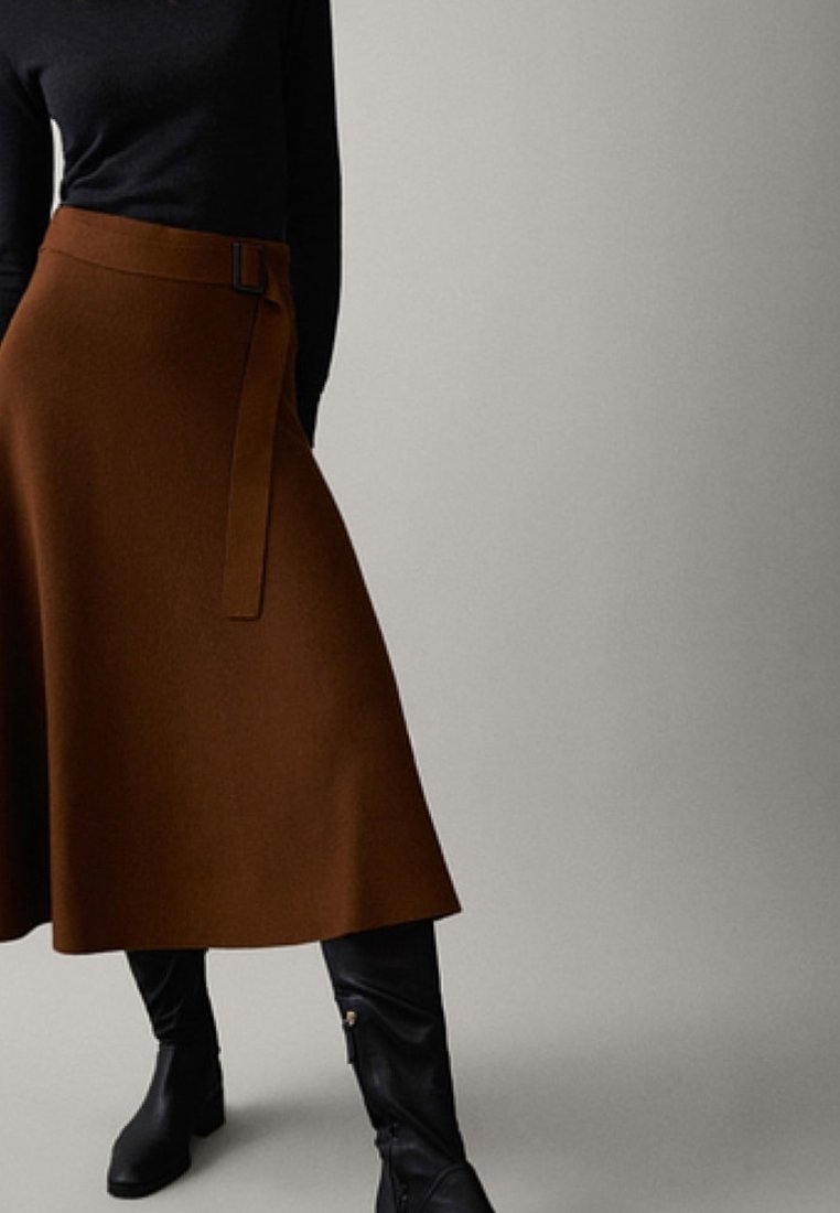 Femme portant une jupe midi marron avec ceinture, un haut noir et des bottes noires montant jusqu'aux genoux, debout devant un fond clair uni.