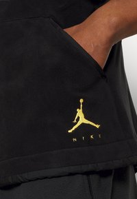 Černá fleecová mikina s kapucí a kapsou typu klokánek. Zlaté logo Jumpman s nápisem "NIKE" v dolní pravé části. Měkký materiál, volný střih.