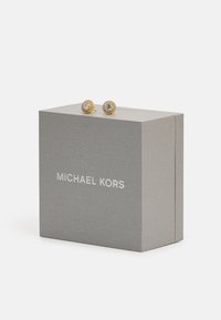Goldene Ohrstecker mit rundem Design, die einen zentralen Edelstein haben, der von kleineren funkelnden Steinen umgeben ist, auf einer grauen Michael Kors Box platziert.