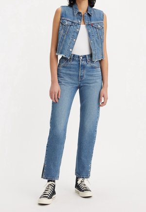 Person iført blå denimvest uden ærmer, højtaljede straight-leg blå jeans med stjernedetaljer ved sømmene, sorte høje sneakers.