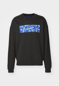 Sweat à manches longues noir avec poignets et ourlet côtelés, avec un patch rectangulaire bleu présentant le texte blanc "HUGO" répété sur la poitrine.