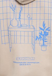 Beige hoodie featuring a blue line drawing of a table with a lamp, vases, and a plant, set against a grid pattern background.