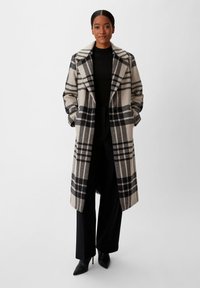 Karierter Mantel in Beige und Schwarz mit Reverskragen, seitlichen Taschen und lockerer Passform, kombiniert mit schwarzen Hosen und hohen Schuhen.