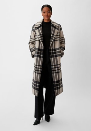 Karierter Mantel in Beige und Schwarz mit Reverskragen, seitlichen Taschen und lockerer Passform, kombiniert mit schwarzen Hosen und hohen Schuhen.