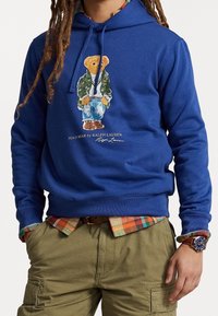 Blå huvtröja i bomull med en grafik av en nallebjörn i jacka och jeans. Kompletterad med en rutig skjorta under och khakifärgade byxor.