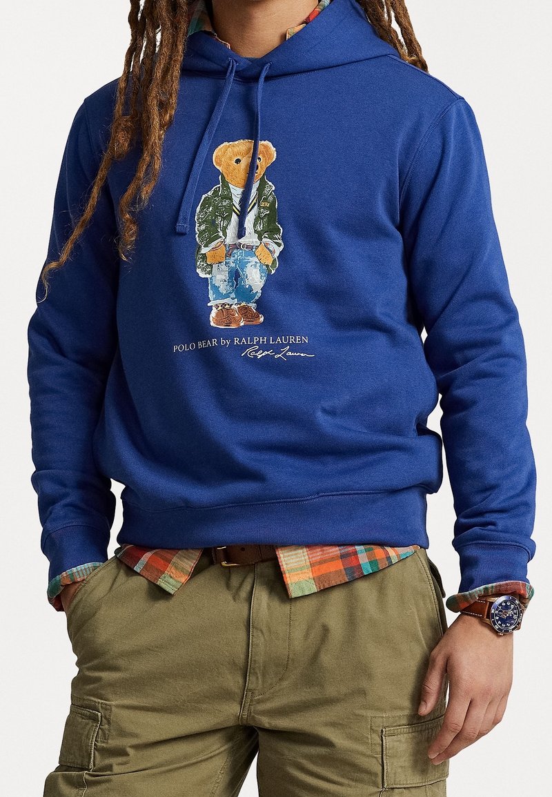 Blå huvtröja i bomull med en grafik av en nallebjörn i jacka och jeans. Kompletterad med en rutig skjorta under och khakifärgade byxor.