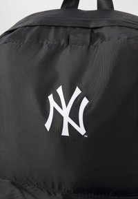 Zaino in nylon nero con un grande logo bianco "NY" sul davanti. Il tessuto ha una finitura liscia e testurizzata con un leggero luccichio.