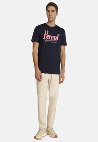 Mørkeblå t-shirt med "Petrol Industries" logo i pink, kombineret med lyse beige bukser og beige sneakers, der viser et afslappet look.
