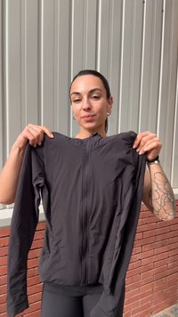 Kvinde med tatoveret arm holder en sort lynlåstrøje op foran en grå panelvæg og en rød murstensfod.