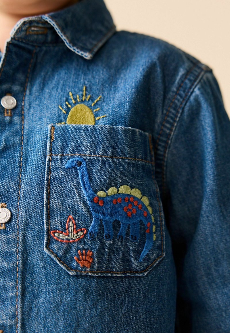Lomme på denimskjorte broderet med en blå dinosaur, orange og grønne pletter, planter og en gul sol delvist synlig over lommen.