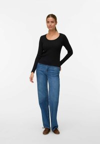 Maglietta nera a maniche lunghe con scollo a barca, abbinata a jeans a gamba larga blu e scarpe marroni, su uno sfondo bianco.