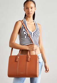 Femme portant un haut à rayures et un jean clair, tenant un grand sac à main en cuir marron avec des poignées et une bandoulière.