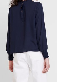 Blouse bleu marine à manches longues, col montant, fermeture par lien au dos et poignets smockés, en tissu lisse, associée à un pantalon blanc.