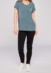 Tyrkysové tričko s krátkým rukávem a kulatým výstřihem, spárované s černými slim-fit kalhotami a bílými teniskami. Na tričku je malá značka.