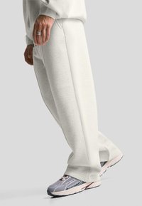 Svetlo sivi trenirka s sproščenim krojem, ki vključuje žepe in elastične manšete, v kombinaciji s sivimi in belimi športnimi copati. Gladka tekstura.