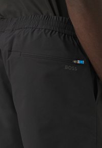 Pantalones cortos ligeros negros con cintura elástica, con un bolsillo con cremallera y el logo "BOSS" en un estampado sutil. Textura suave.