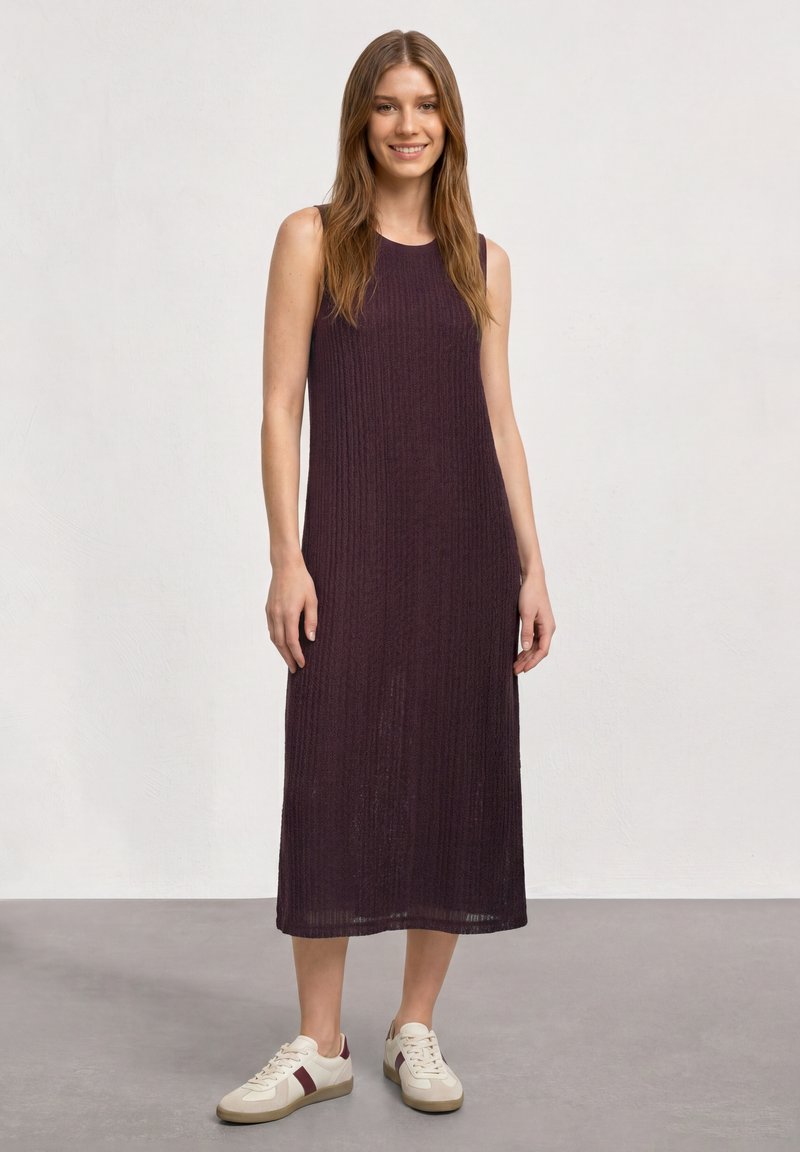 Jeune femme debout souriante, portant une robe midi sans manches côtelée de couleur violet foncé et des baskets blanches avec des rayures bordeaux.